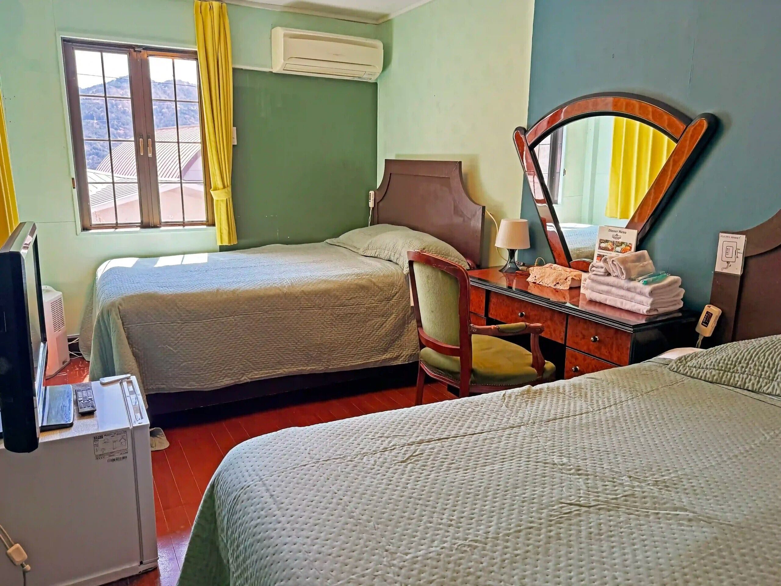 A hotel room with two beds, a desk with a mirror, a chair, a small table with towels, and an air conditioning unit.