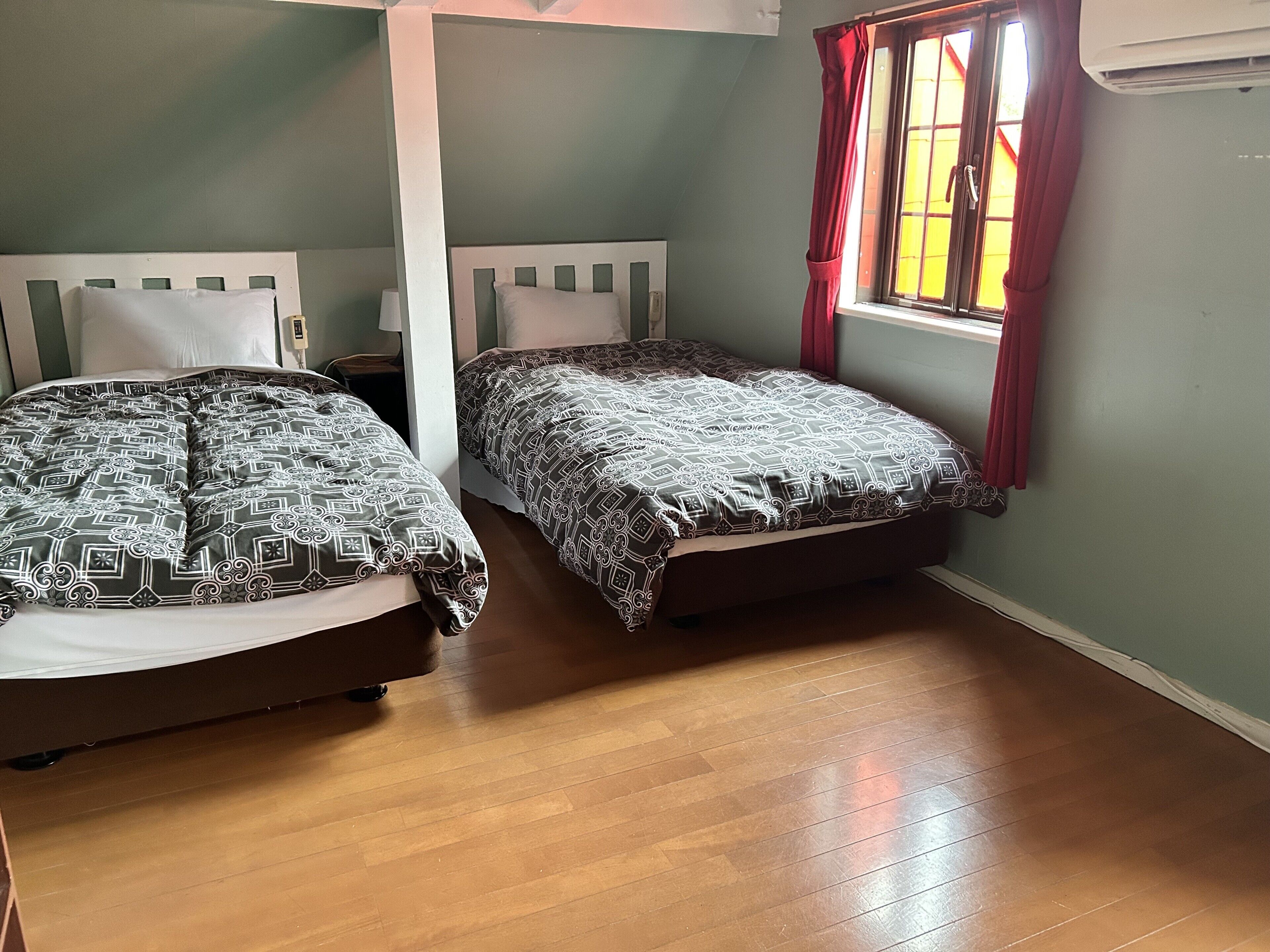 Two beds with patterned bedding, a wooden floor, a window with red curtains, and an air conditioning unit.