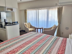 A hotel room with a bed, two armchairs, a small table, a kitchenette, and a large window with curtains.