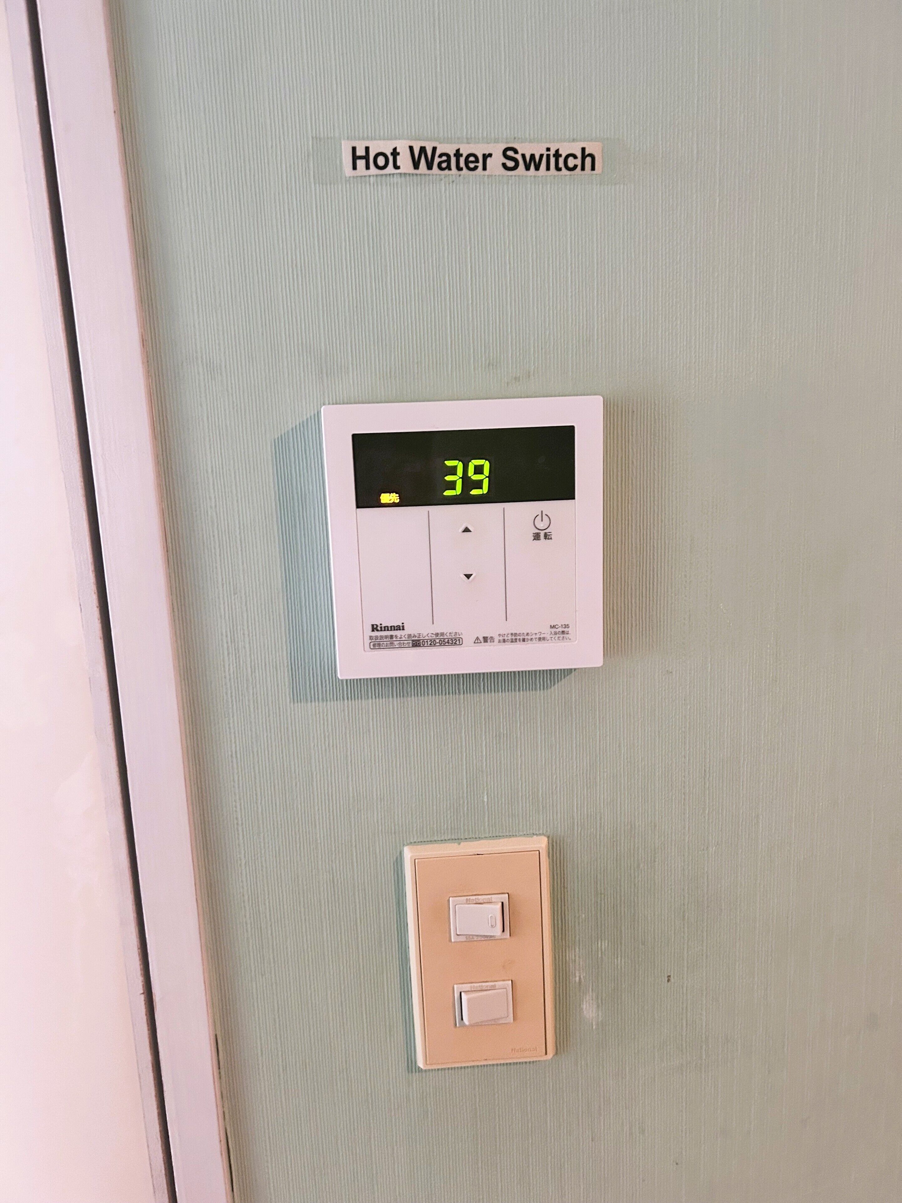 Interruptor de agua caliente con pantalla digital que indica 39 grados Celsius, panel de control y un interruptor de luz estándar debajo.