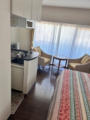 Ein Hotelzimmer mit Bett, einer kleinen Küchenzeile, zwei Sesseln, einem runden Tisch und Blick durch leichte Vorhänge.