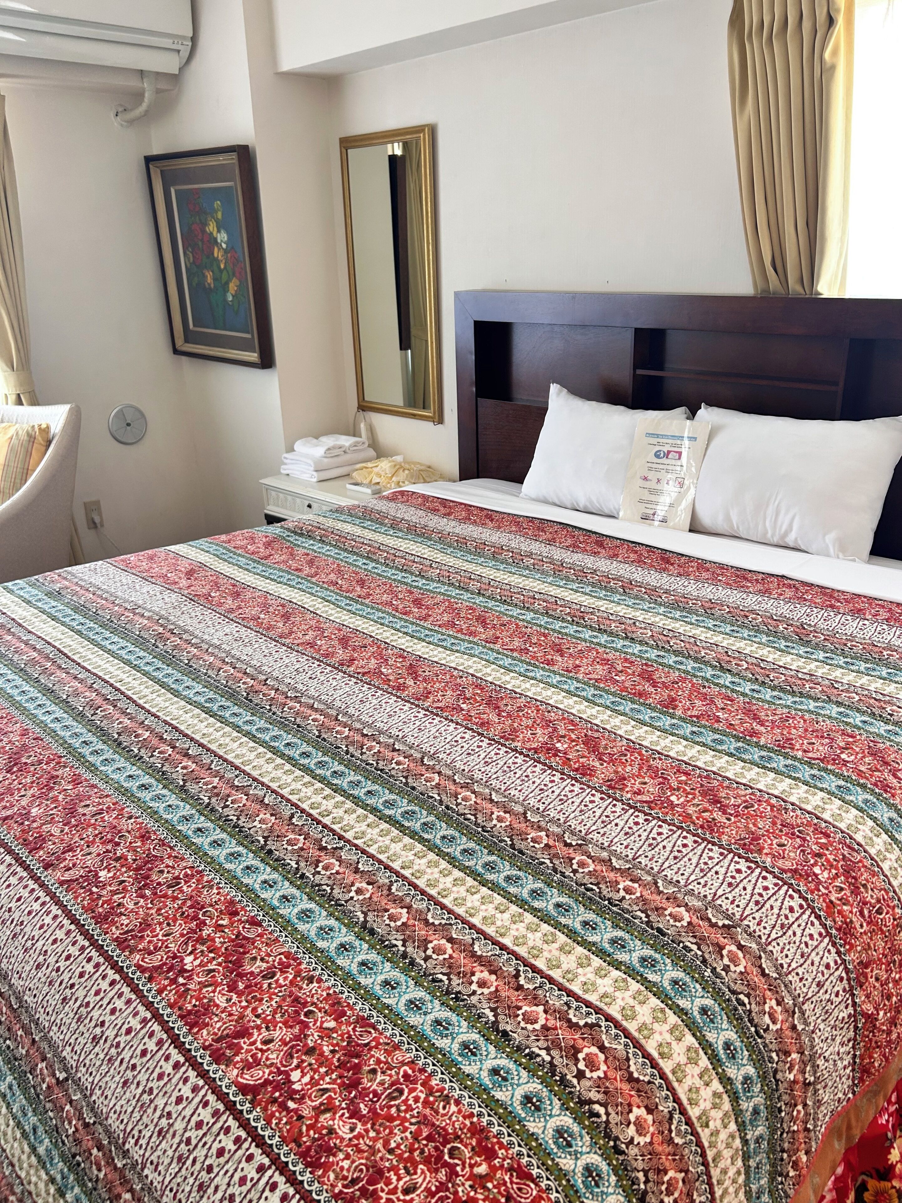 A hotel room with a large bed, a headboard, a bedside table with a lamp, a clock, and a framed picture on the wall.