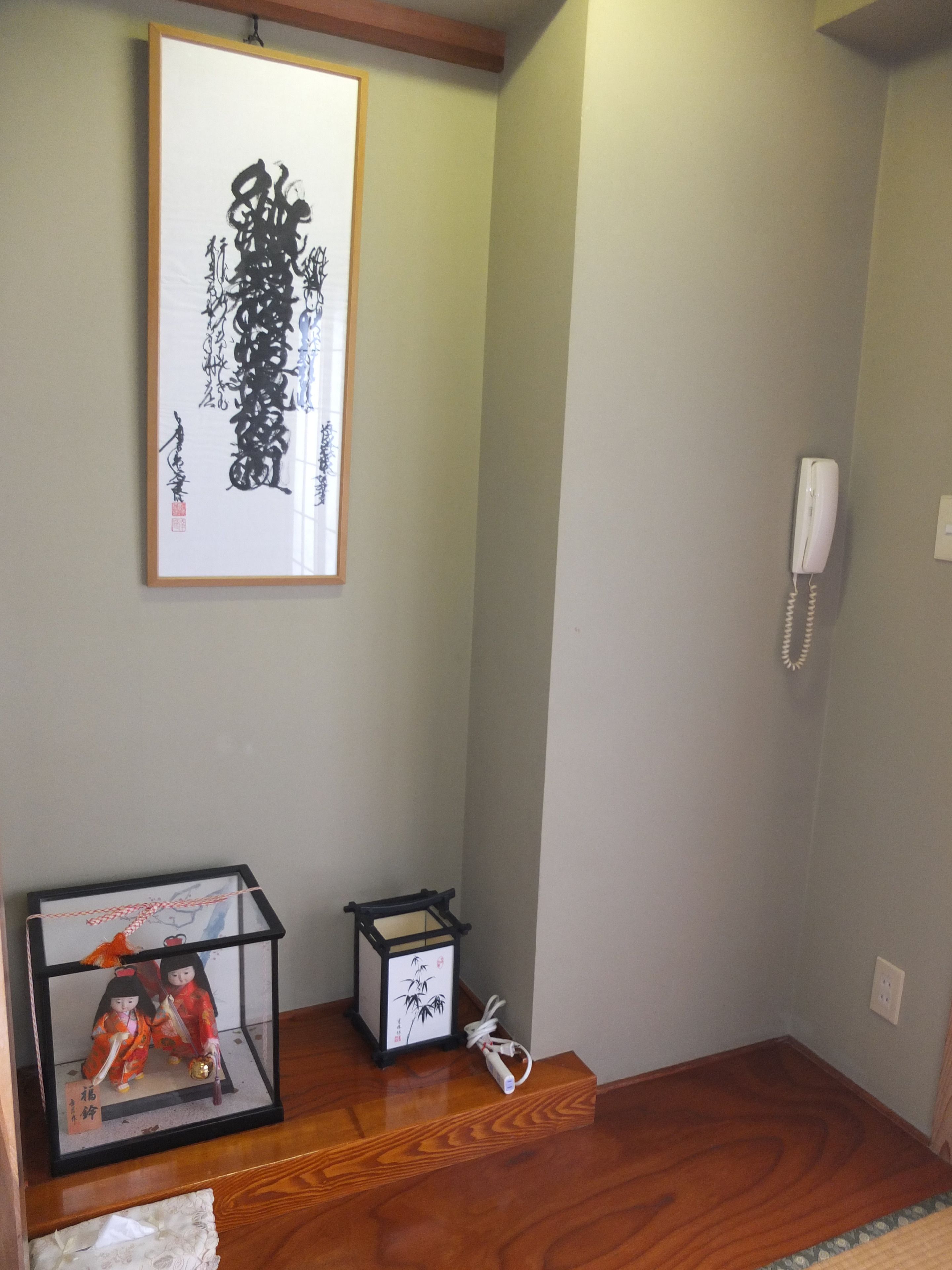 Ein Zimmer mit einem gerahmten Kalligraphie-Kunstwerk, einer traditionellen japanischen Puppenausstellung, einem kleinen Tisch und einem Telefon an der Wand.
