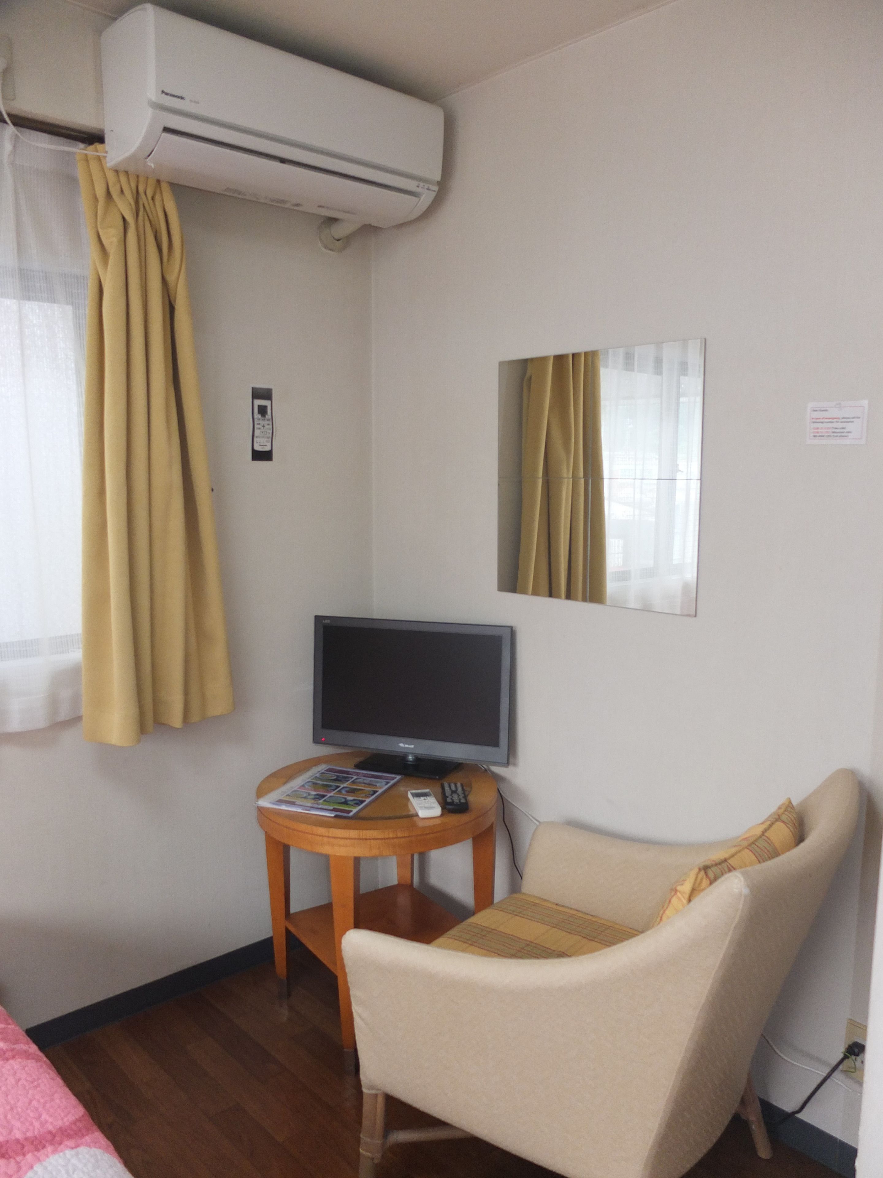 Ein Hotelzimmer mit einem Flachbildfernseher auf einem Holztischchen, einem beigen Sessel, einer Klimaanlage und einem Fenster mit gelben Vorhängen.