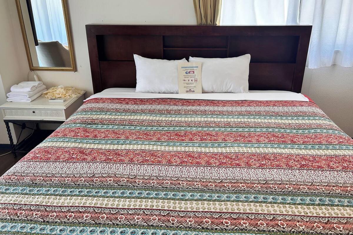 A hotel room with a large bed, a bedside table with a lamp, a mirror, and a sign with room amenities.