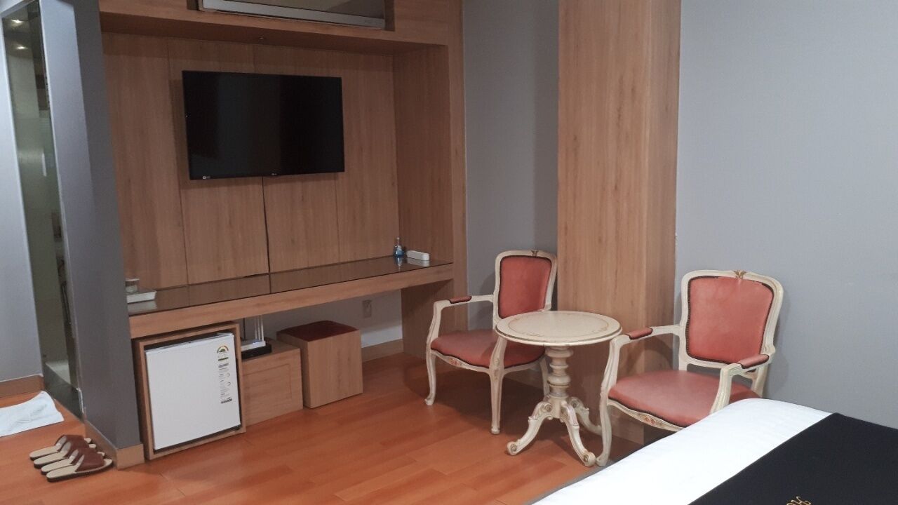 A hotel room with a wooden cabinet, a flat-screen TV, a small refrigerator, a round table with two chairs, and a bed.