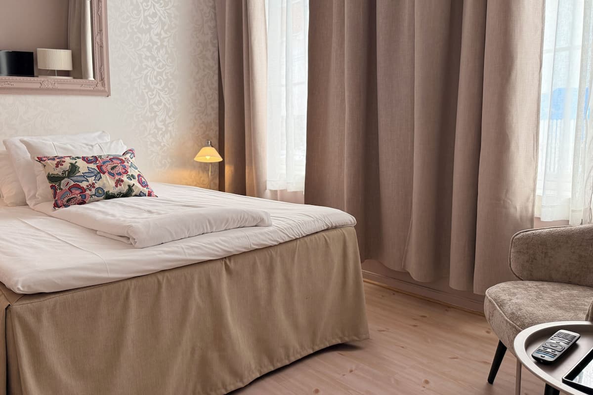 Una cama bien hecha con un cojín floral, una lámpara de noche y una silla con un control remoto sobre un piso de madera.