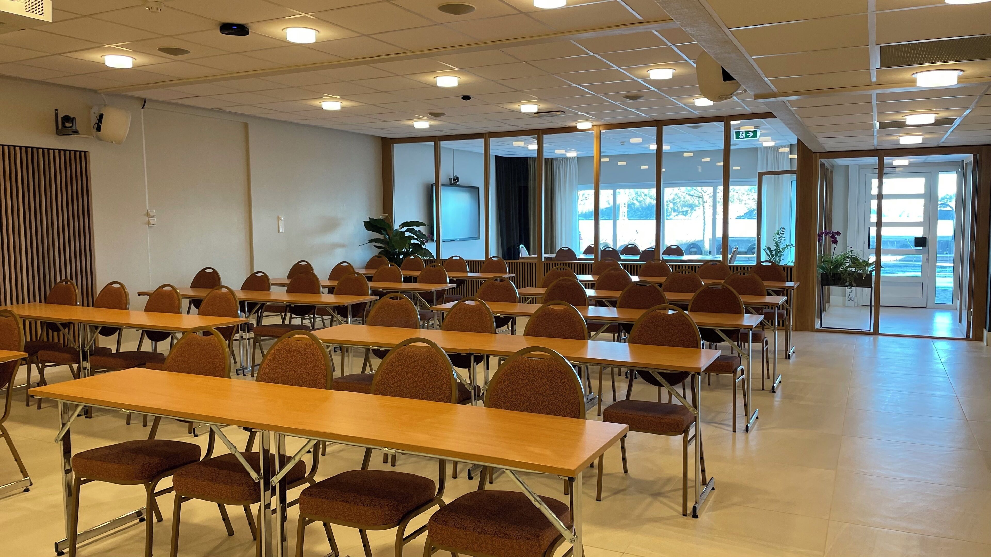 Un salón de conferencias con varias mesas y sillas, amplias ventanas y una puerta que da al exterior.