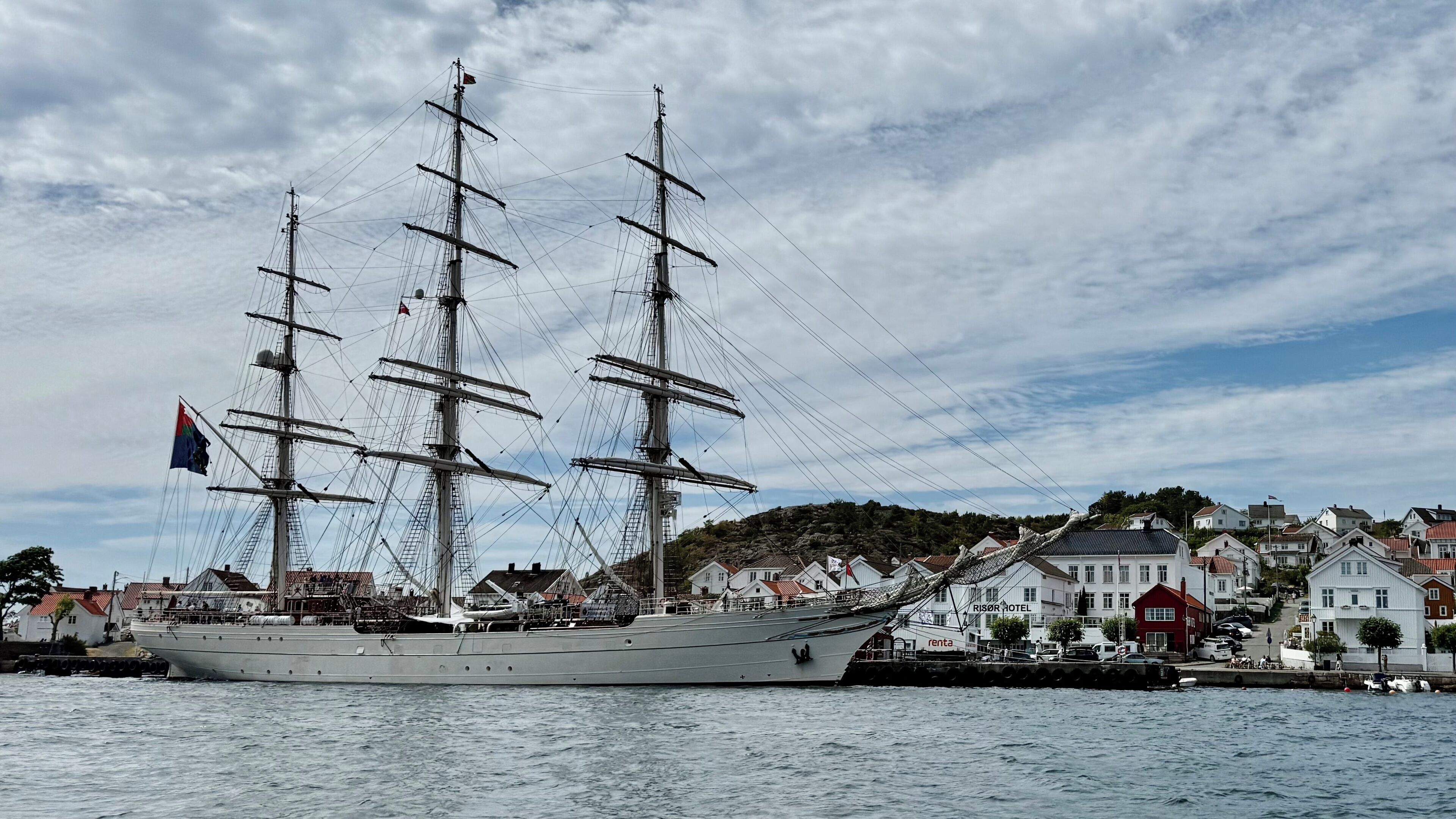 A large sailing ship docked at a harbor with a row of houses in the background.