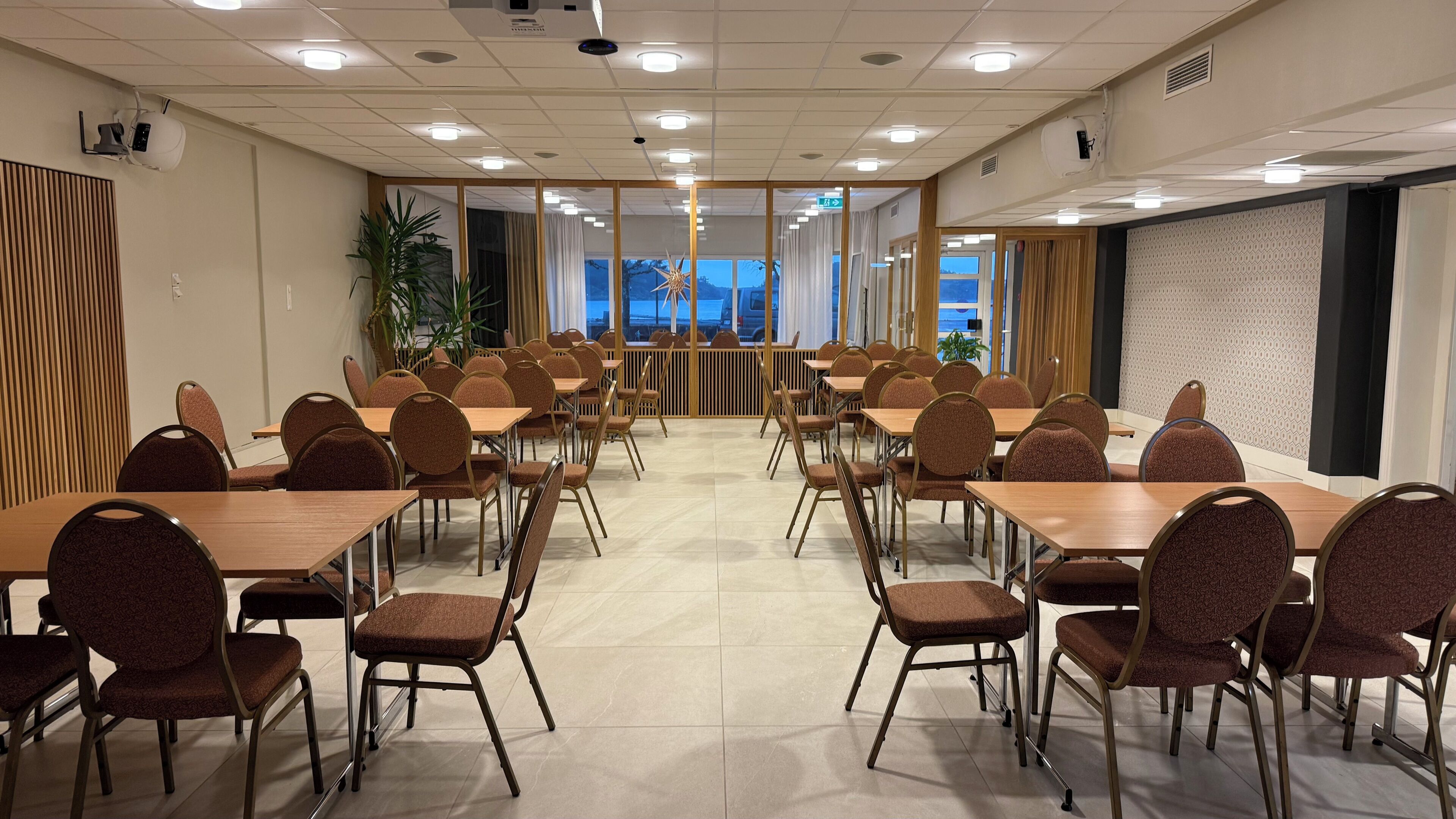 Une salle de conférence dotée de plusieurs tables et chaises, d’une cloison en bois et offrant une vue sur l’extérieur grâce à de grandes fenêtres.
