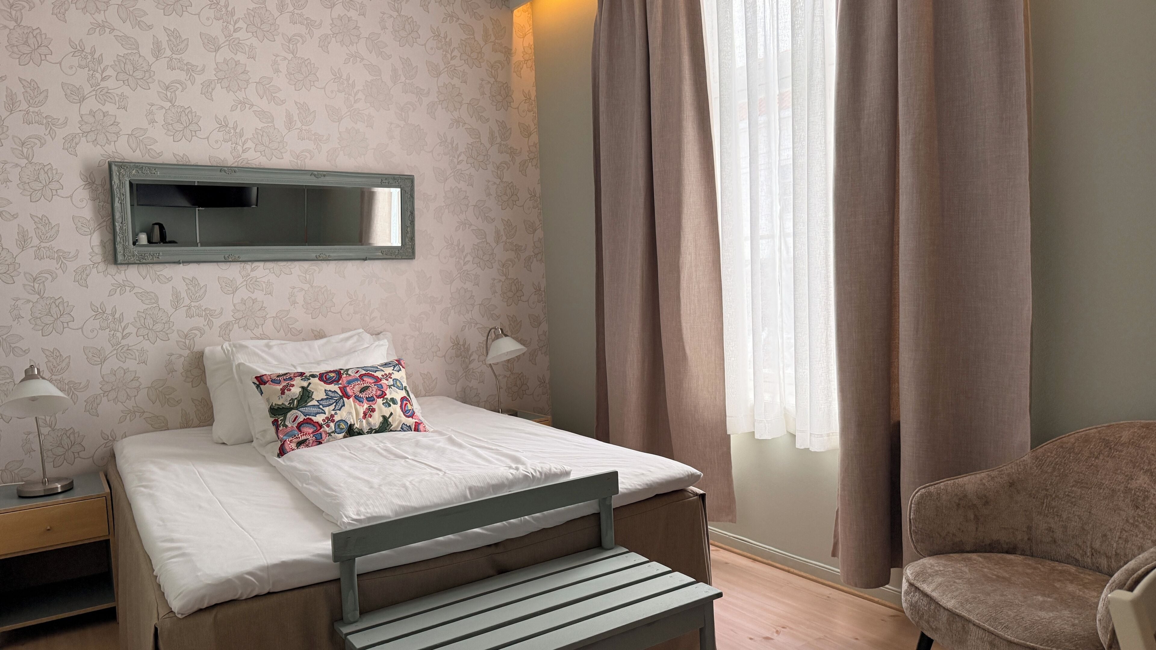 A bedroom with a bed, a nightstand, a lamp, a chair, a mirror, and a window with curtains.