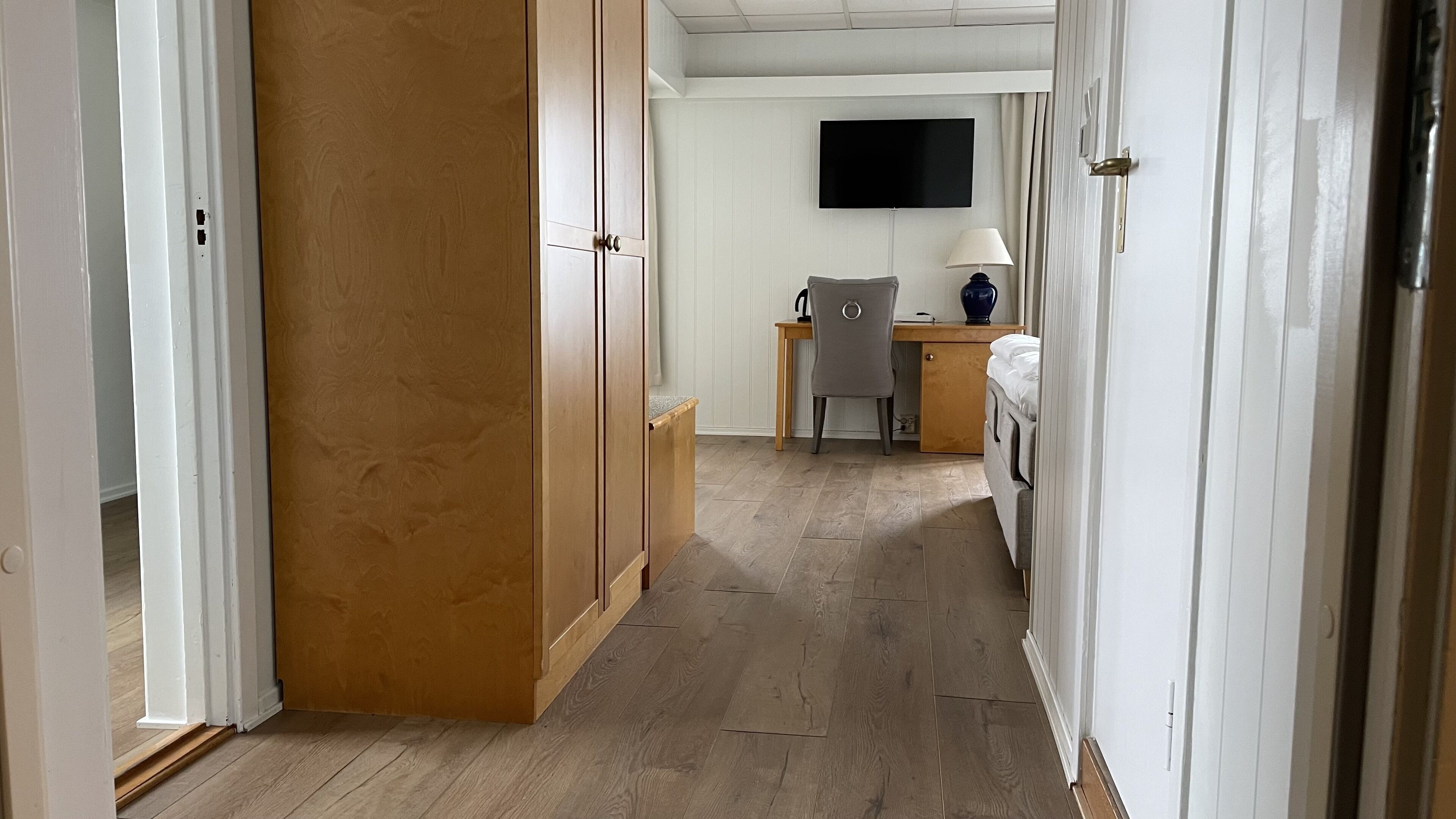 Un pasillo con suelo de madera, un escritorio con silla, una cama y un televisor montado en la pared.
