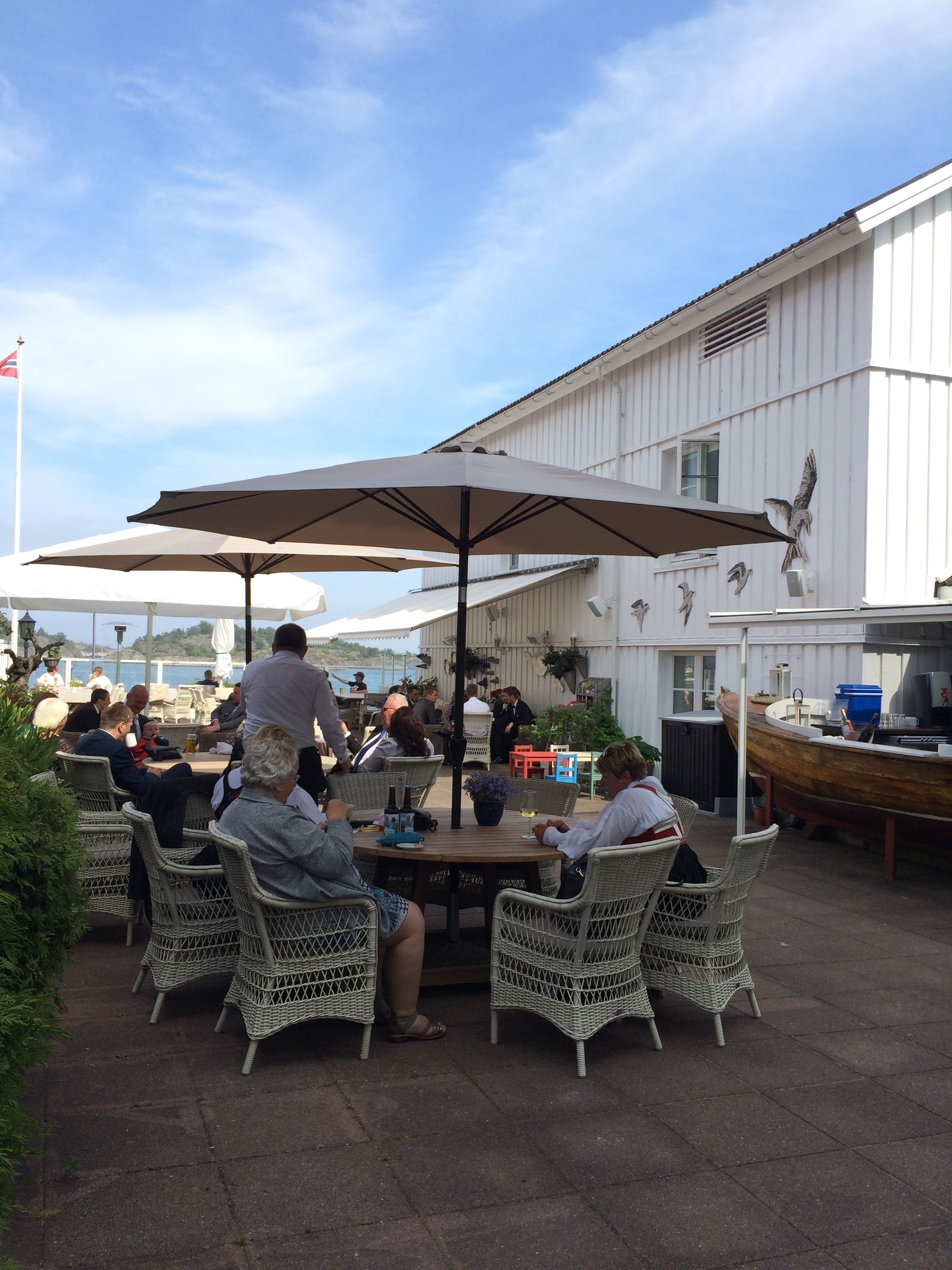 An outdoor dining area with wicker chairs and large umbrellas, a white building with decorative bird sculptures, and a flag in the background.