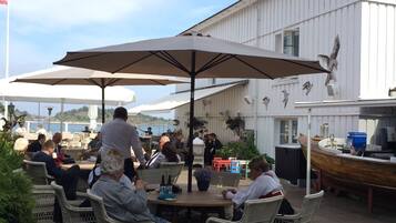 An outdoor dining area with wicker chairs and large umbrellas, a white building with decorative bird sculptures, and a flag in the background.
