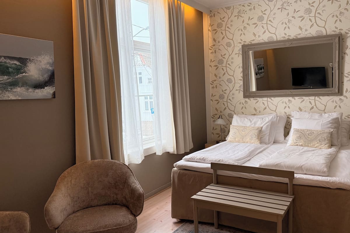 Habitación de hotel con cama, una silla, escritorio, lámpara, espejo y ventana con cortinas.