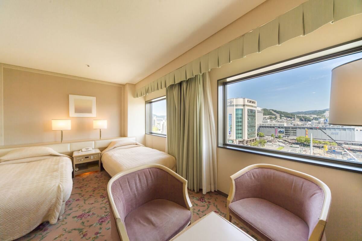 Une chambre d’hôtel avec deux lits, deux tables de chevet, une fenêtre donnant sur un paysage urbain et une petite table avec une chaise.