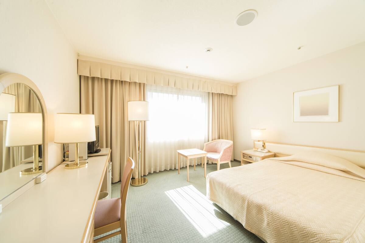 Une chambre d’hôtel comprenant un lit, un bureau avec une chaise, une table de chevet avec une lampe, un miroir et une fenêtre avec des rideaux.