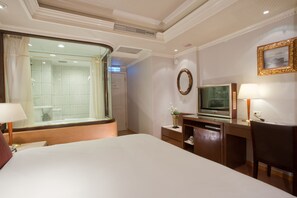 Habitación de hotel con una cama grande, televisión, escritorio y baño con cabina de ducha de vidrio.