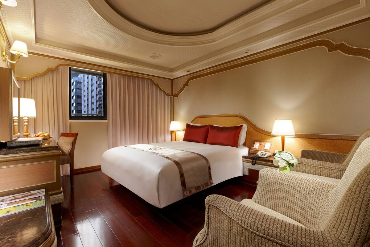 Quarto de hotel com uma cama grande, uma cadeira, uma mesa e uma janela com cortinas.