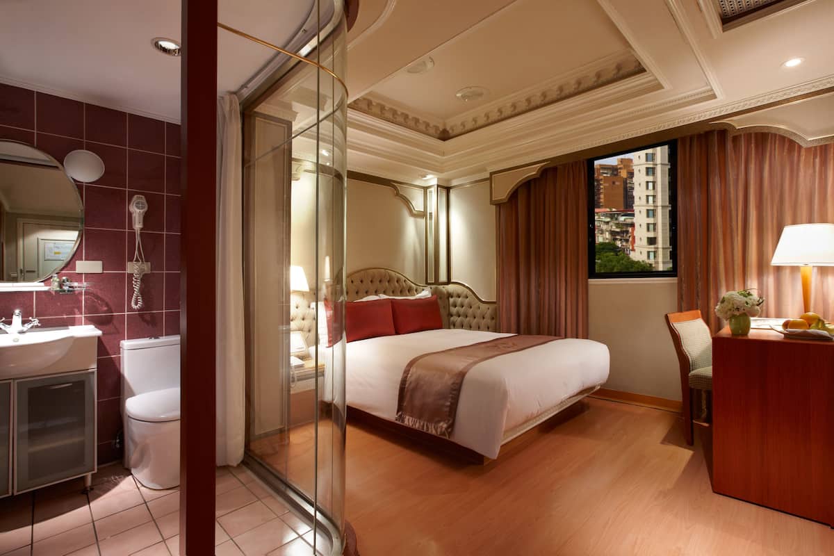 Quarto de hotel com cama, escrivaninha, cadeira, toucador com espelho e banheiro com vaso sanitário e pia.