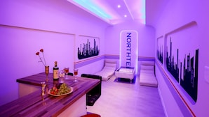 Una habitación moderna con un mural de paisaje urbano, una zona de bar con una botella y vasos, y un sillón reclinable con la etiqueta 'NORTHTHE'.