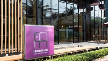 A modern building with large glass windows, a sign for 'Hotel icon Sukhumvit 2 Bangkok', and outdoor seating.