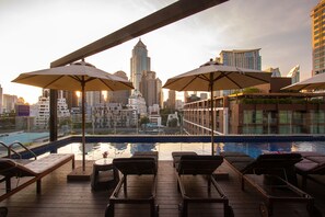 A rooftop pool area with lounge chairs and umbrellas, overlooking a cityscape with modern buildings.