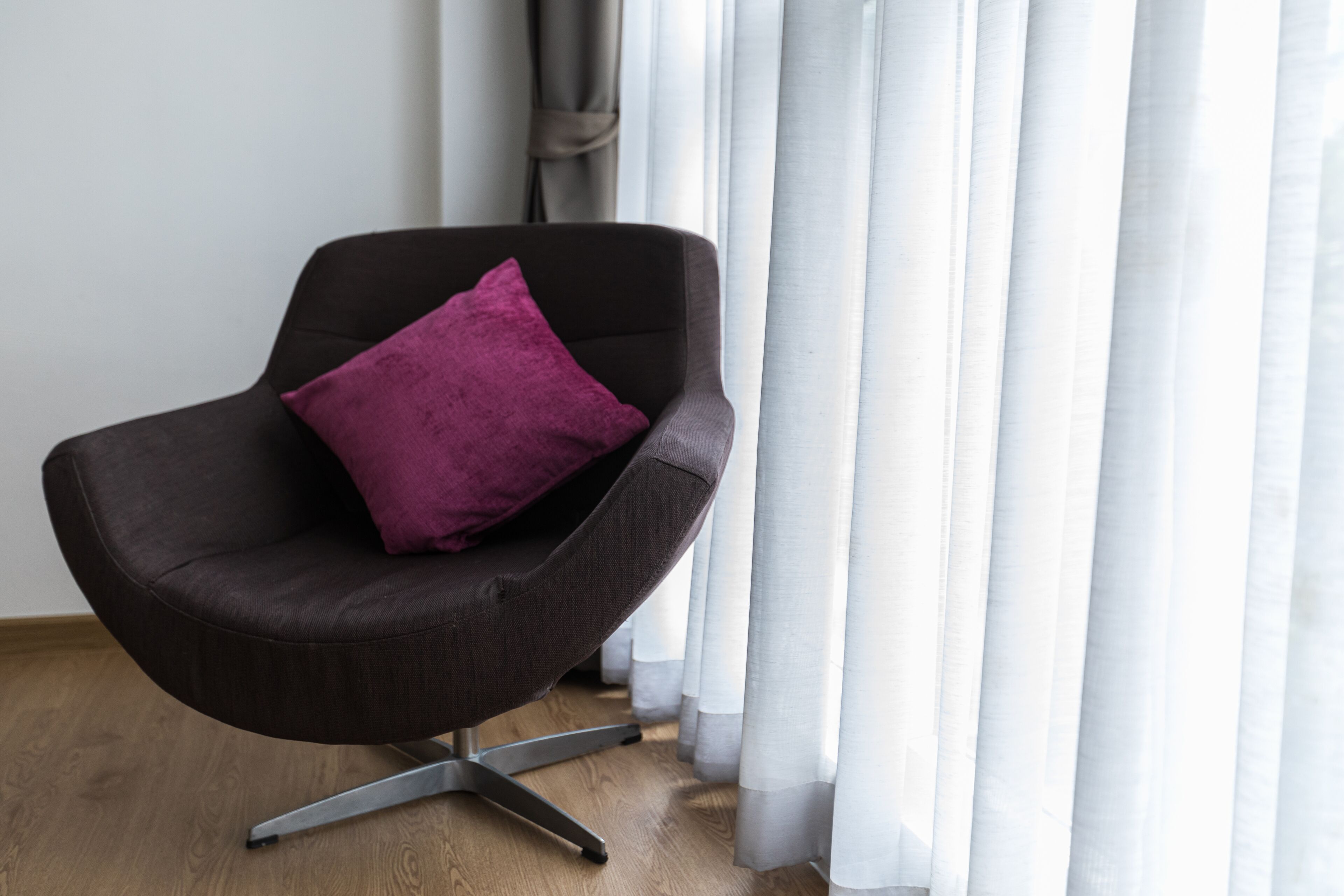 A modern armchair with a metallic base, positioned in front of a window with sheer white curtains.