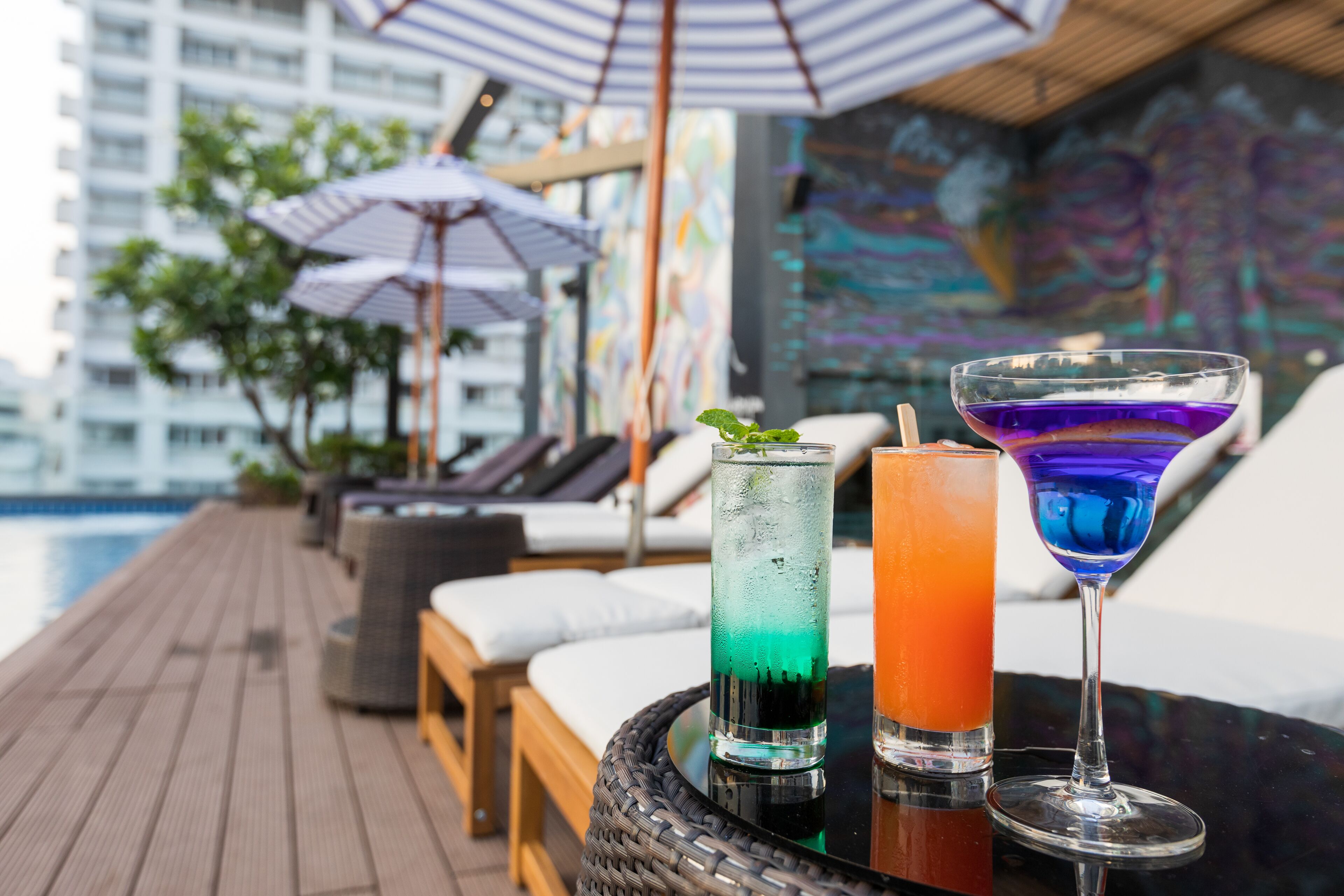 Un espace aménagé au bord de la piscine, avec des chaises longues et des parasols, une table en verre sur laquelle reposent deux boissons, et un paysage urbain en arrière-plan.