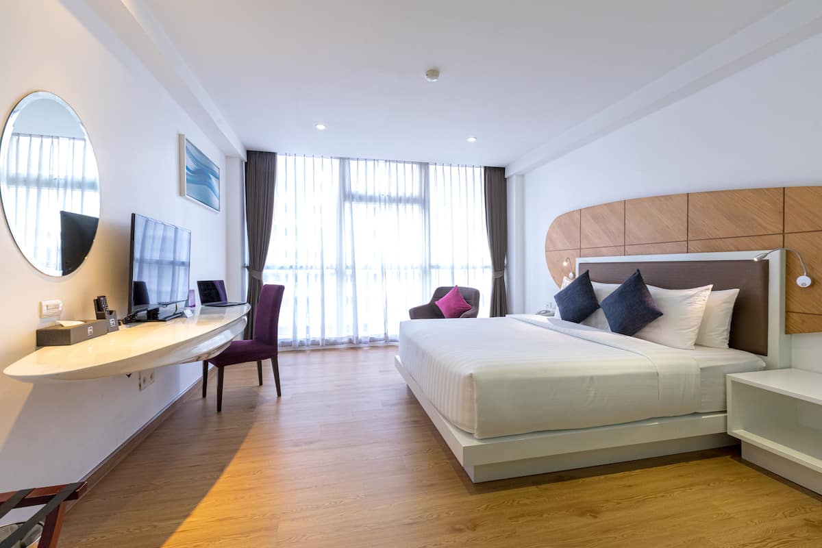 Een hotelkamer met een bed, nachtkastje, bureau, stoel en een groot raam met gordijnen.