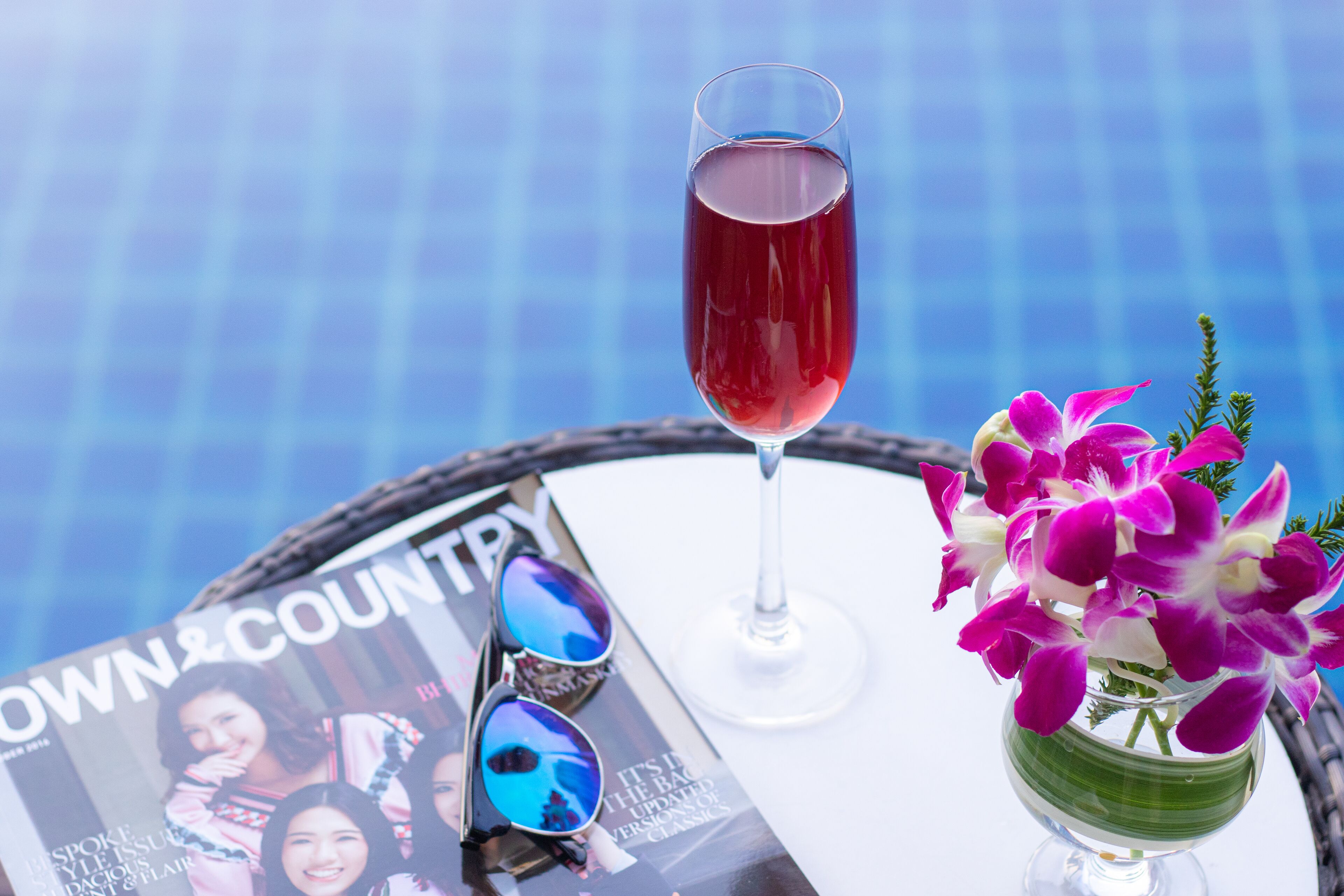 Una revista, un vaso de bebida roja y un florero con flores moradas sobre una mesa junto a una piscina.