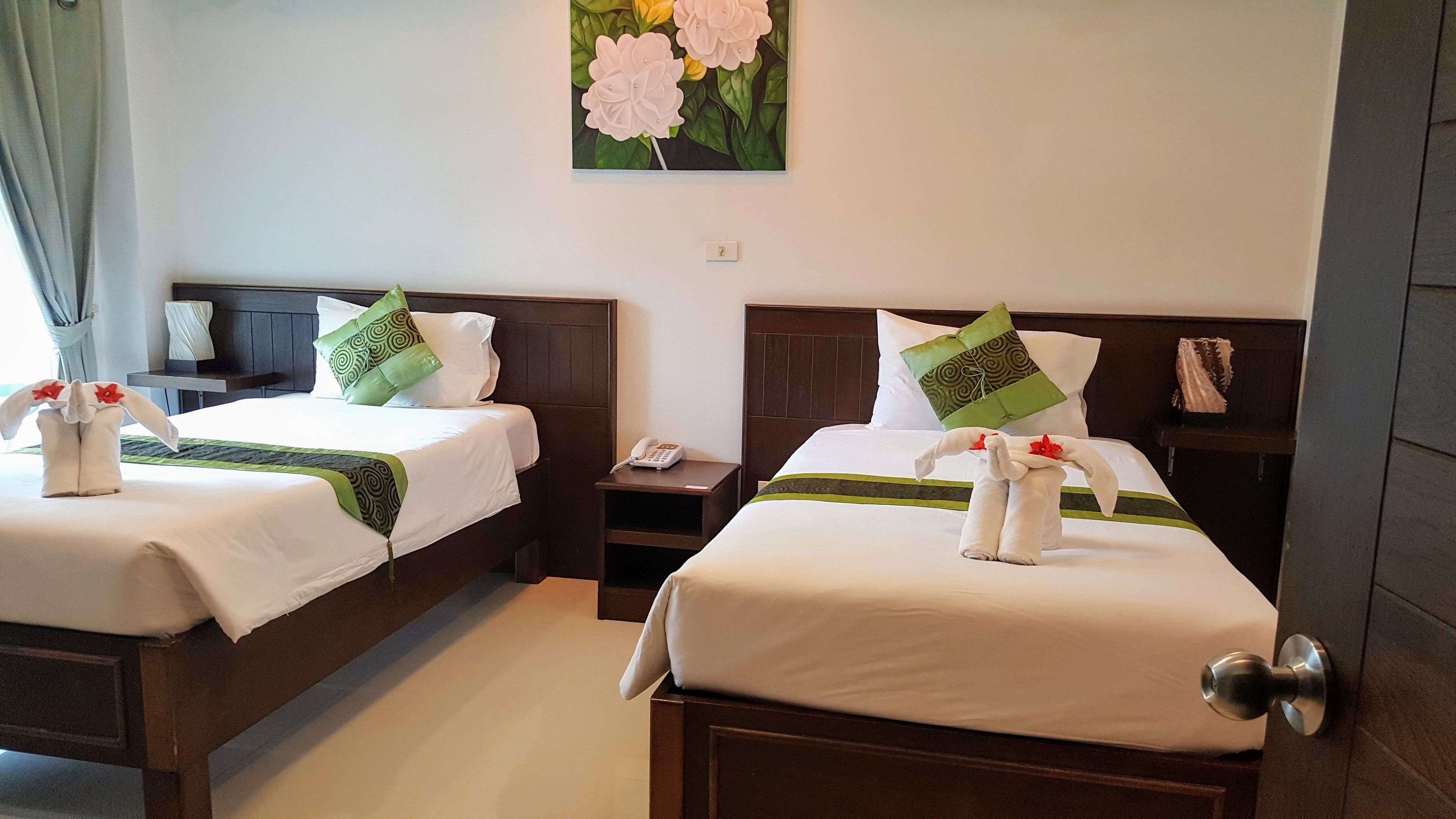 Two neatly made beds with white linens, green and patterned pillows, and towels arranged as a decorative element.