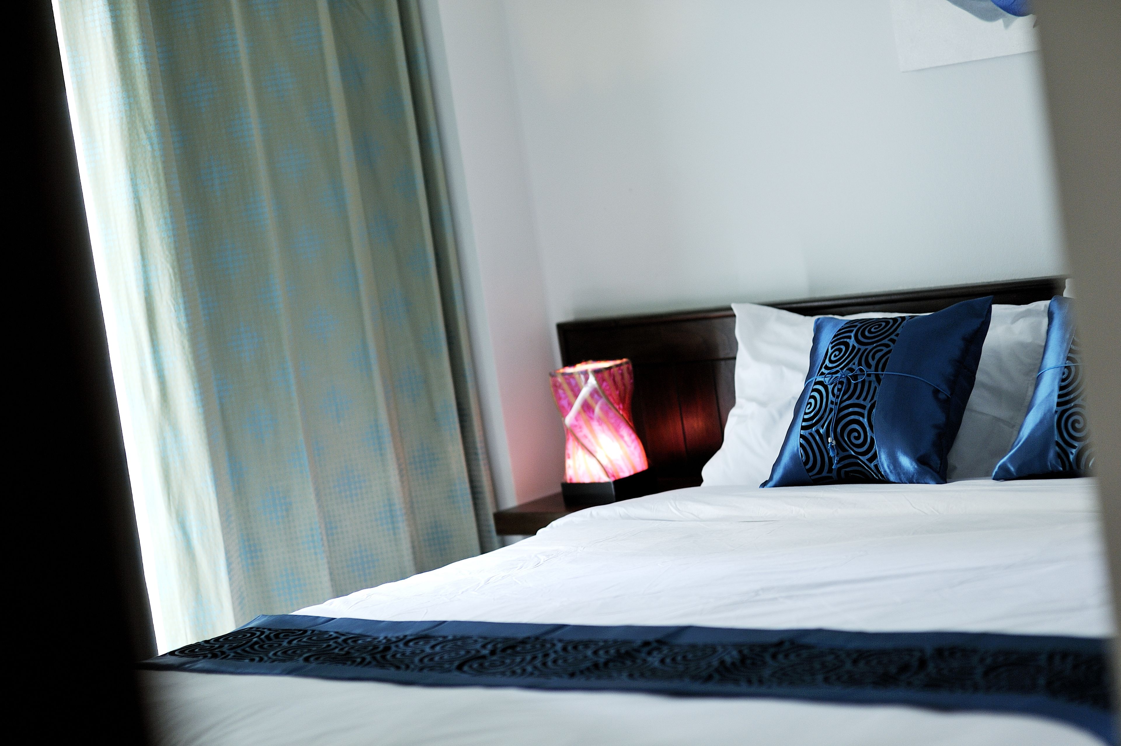 Habitación de hotel con una cama bien hecha, una mesita de noche con lámpara y una ventana con cortinas.