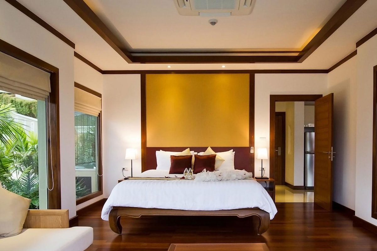 A modern hotel room with a large bed, bedside lamps, a view of greenery, and a kitchen area.