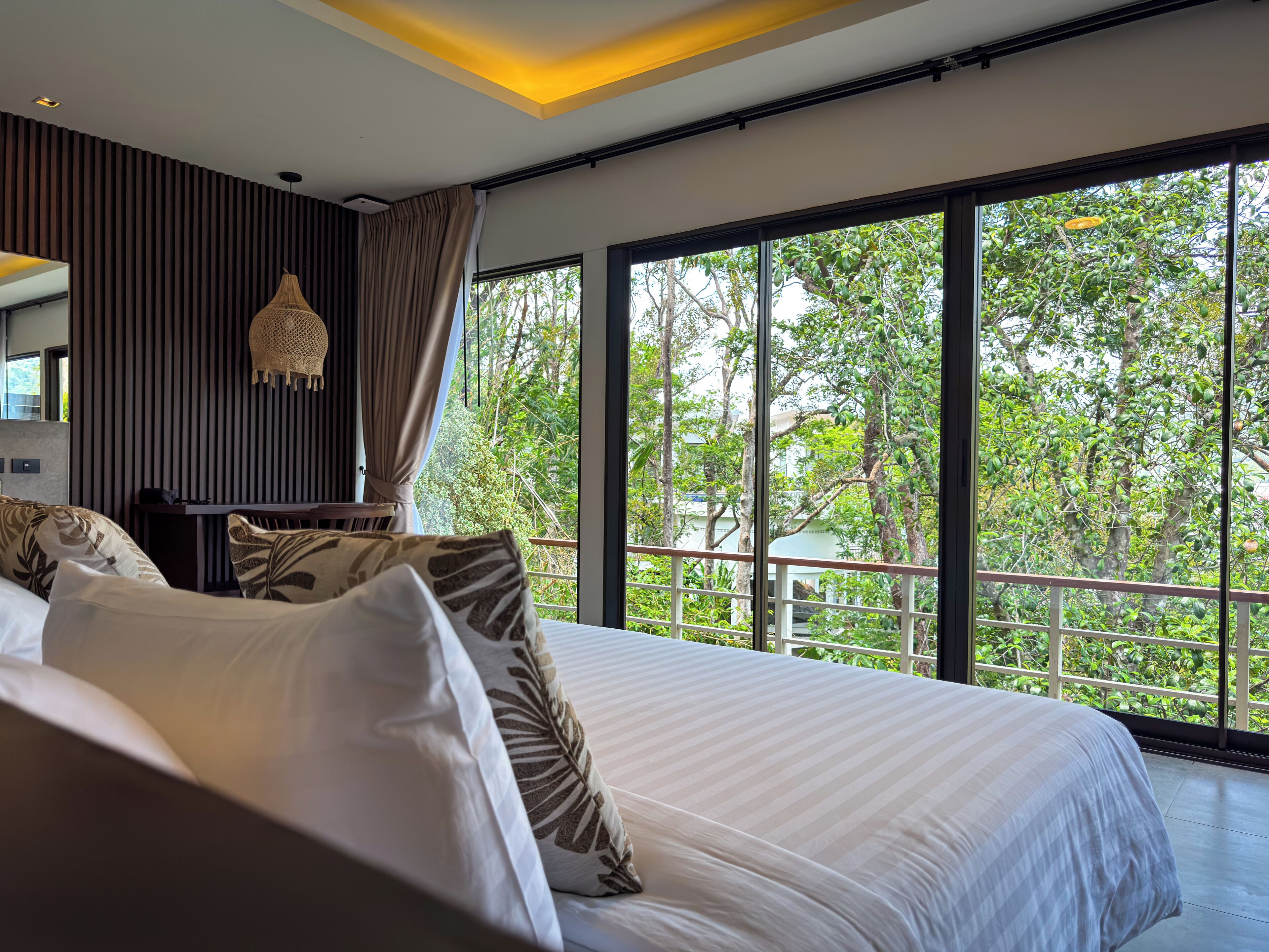 Ein modernes Schlafzimmer mit einem großen Bett, einem Schreibtisch und Blick auf üppiges Grün durch große Fenster.