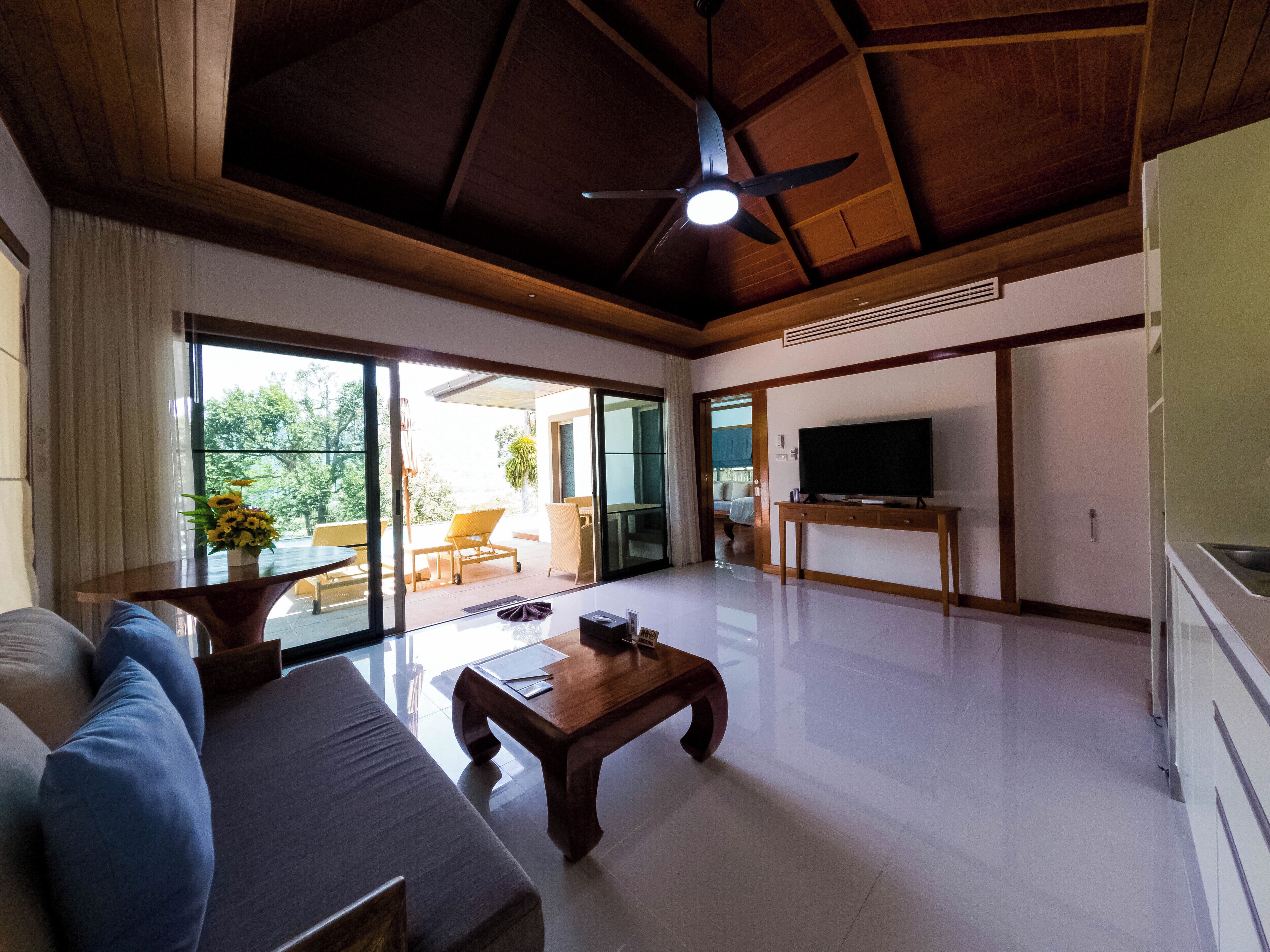 A spacious living room with a ceiling fan, a large TV, and a sliding glass door leading to an outdoor area with a patio and a view of greenery.