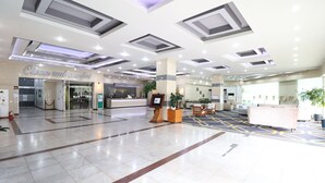 Un vestíbulo de hotel con mostrador de recepción, un letrero que dice 'Limpio y Cómodo', un letrero de Grand Hotel, y una zona de recepción con una planta y asientos.