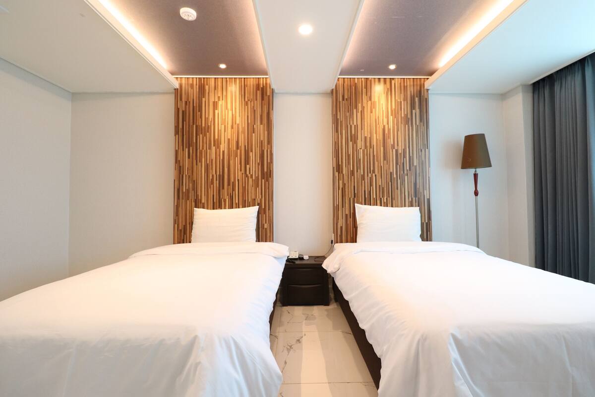Habitación de hotel con dos camas individuales, paneles de madera, una mesita de noche, una lámpara y vistas al exterior.