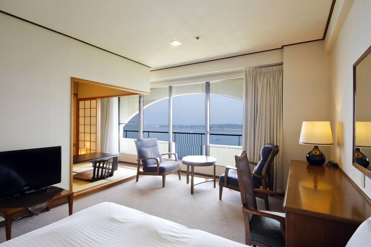 Een hotelkamer met een bed, een televisie, een bureau met een lamp, een stoel, een klein tafeltje en een balkon met uitzicht op zee.