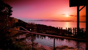 Une vue sur le coucher de soleil depuis un balcon donnant sur une étendue d’eau, avec un pont et des montagnes en arrière-plan.