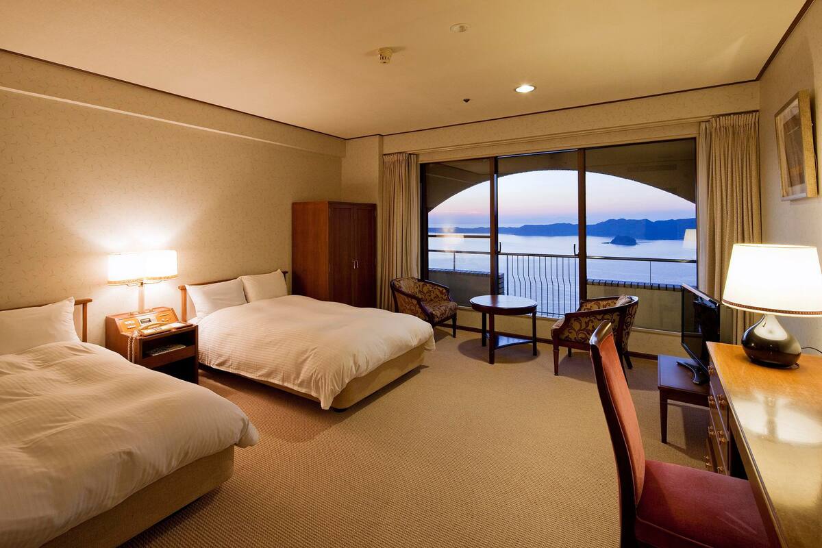 Een hotelkamer met twee bedden, een tafeltje, een stoel en uitzicht op zee door grote ramen.