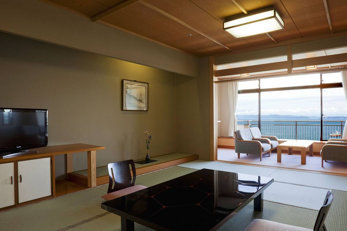 A hotel room with a TV, a dining table, a sofa, and a view of the sea.