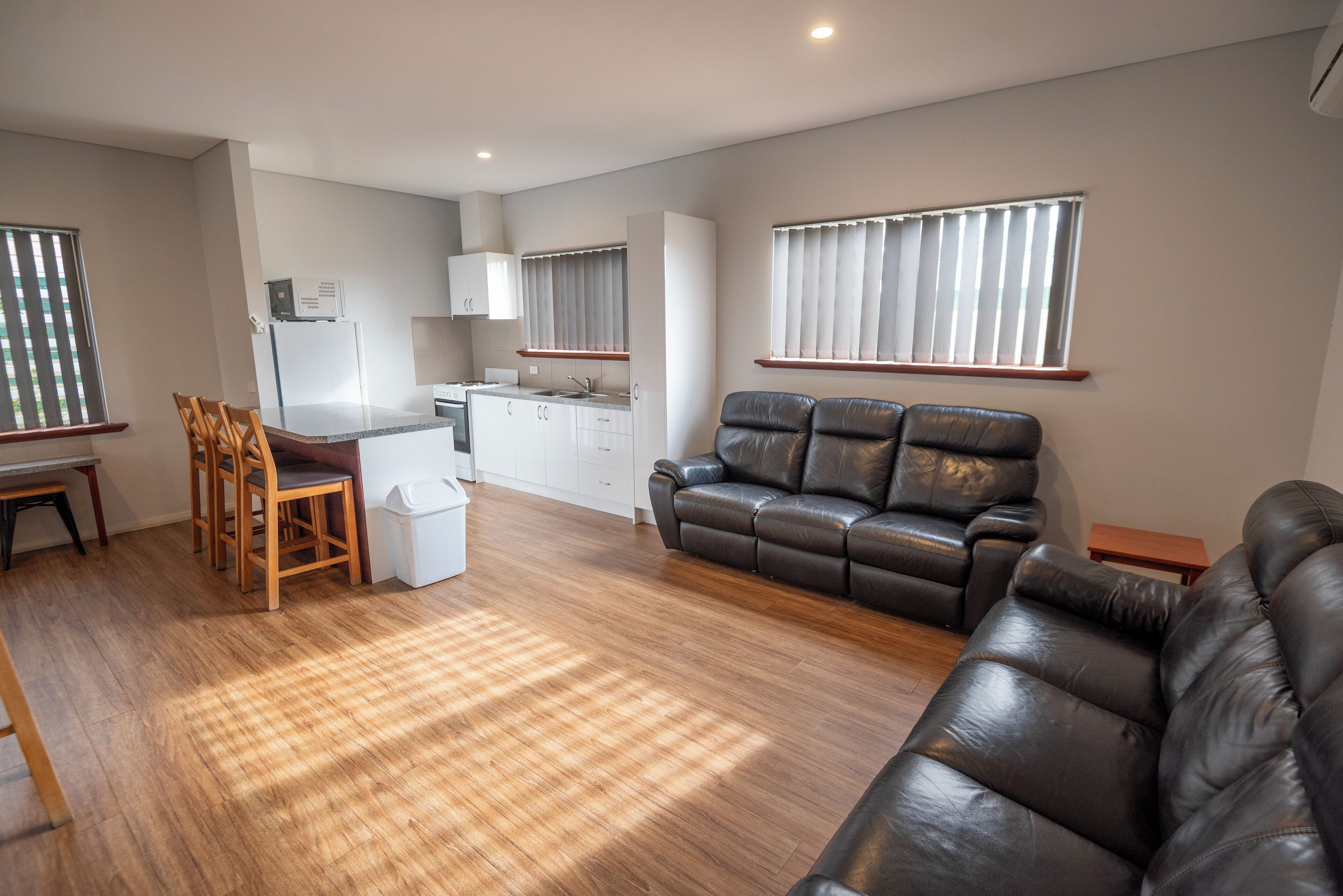 Een moderne woonkamer met een leren bank, een houten eettafel en stoelen. In de achtergrond is een keuken met witte kasten en een koelkast zichtbaar.