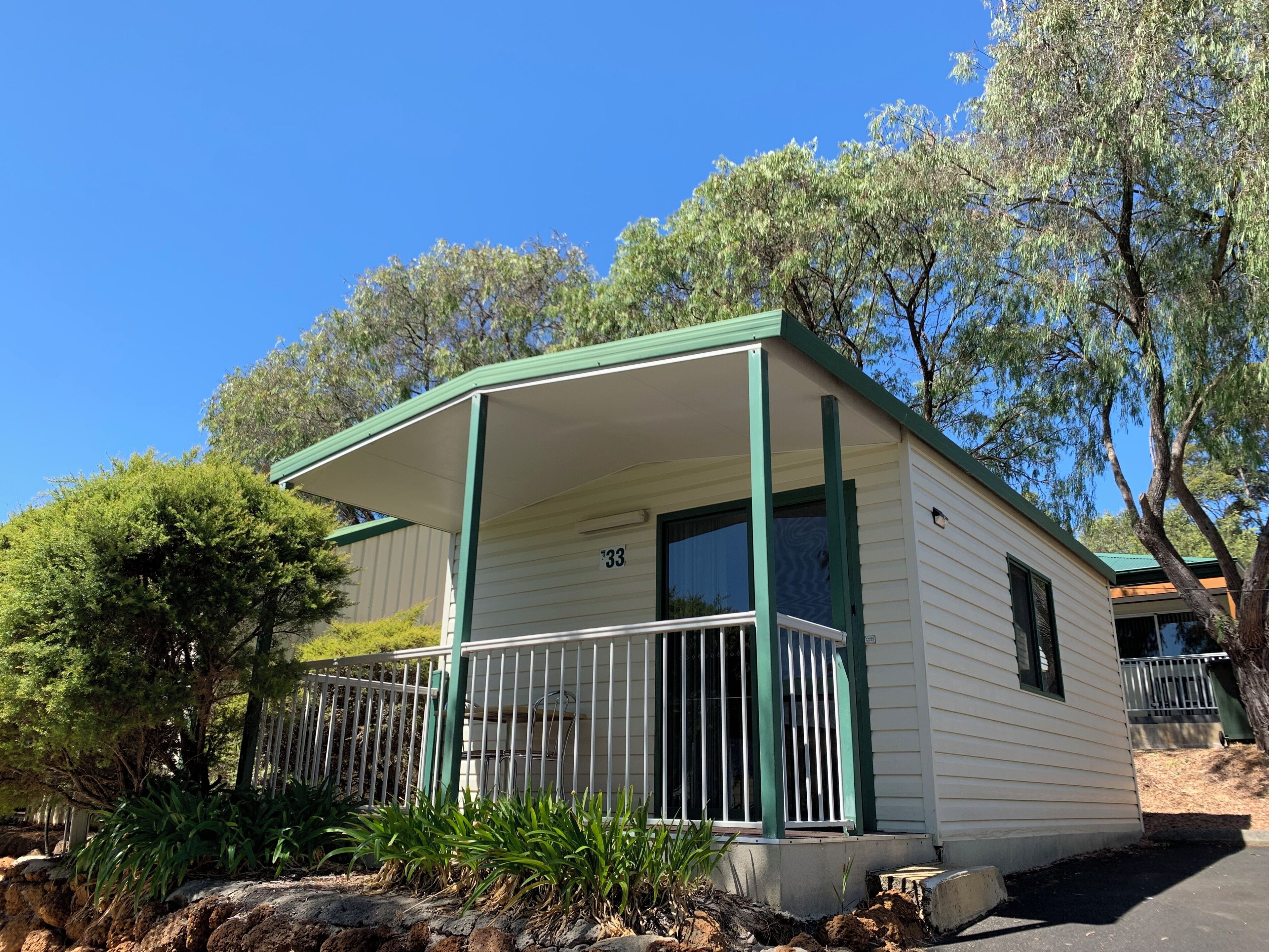 Casa móvel térrea com telhado verde e revestimento branco, número 33, cercada por árvores e um quintal com cerca.