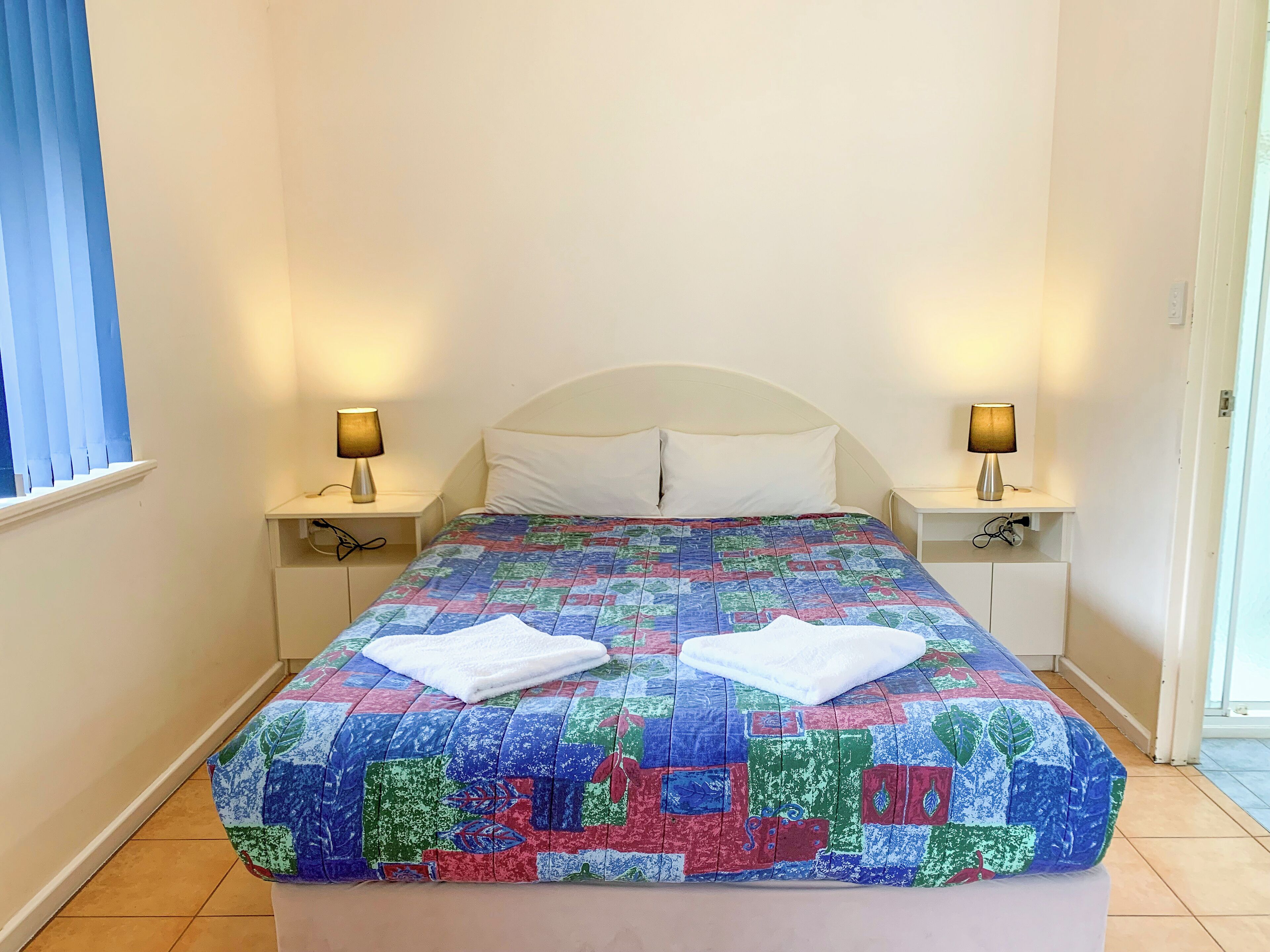 Un letto rifatto con cura, un patchwork colorato, due lampade da comodino e una finestra con persiane.