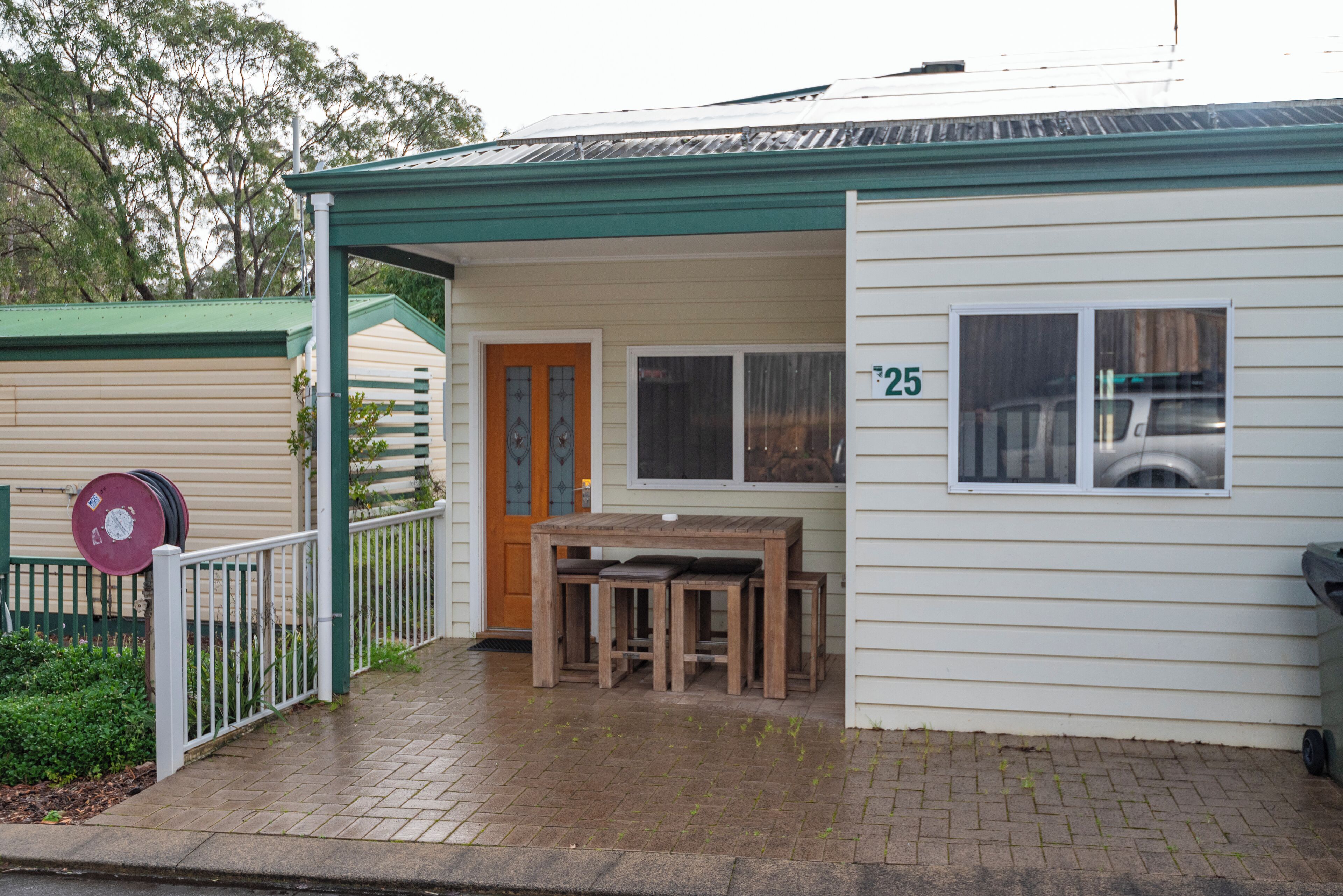 A small, single-story building with a green roof and white siding. It features a number '25' on the side, a wooden table and benches outside, and a white fence with a gate.
