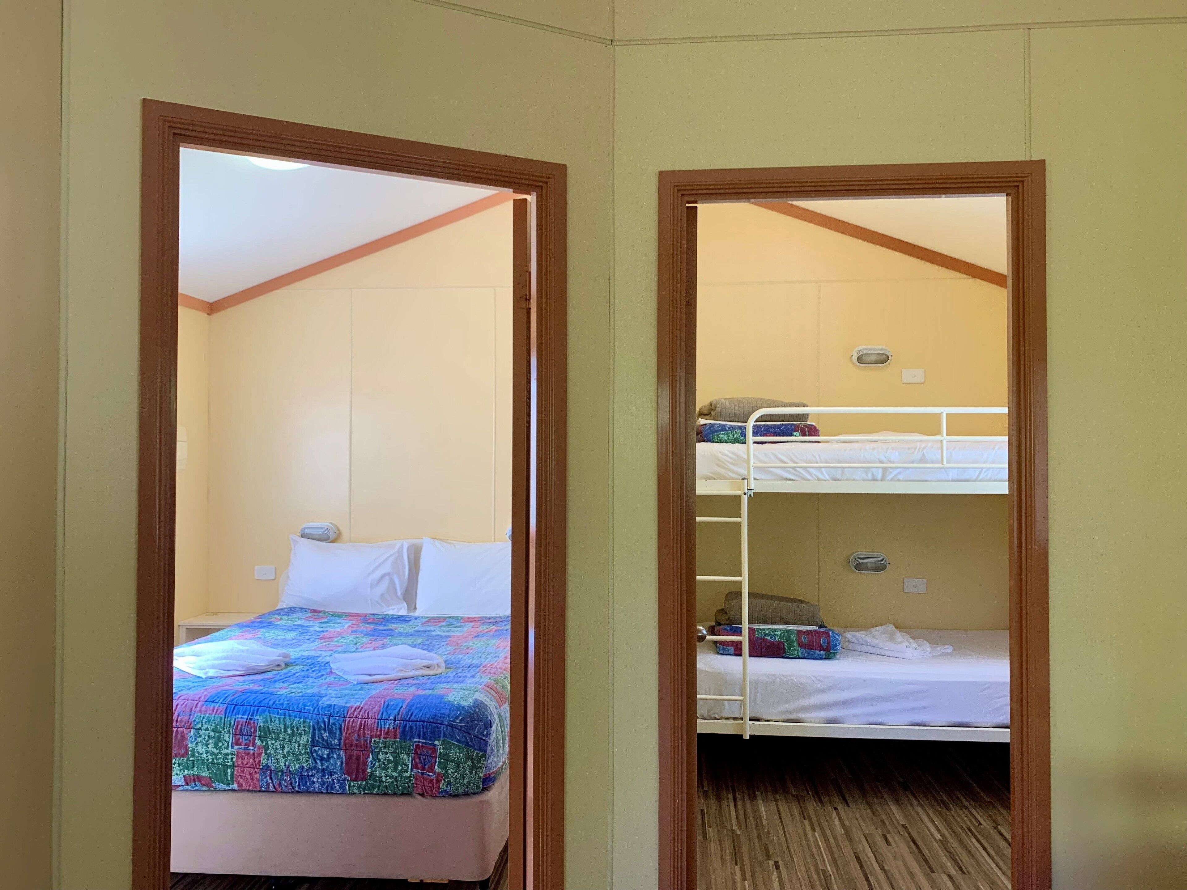A room with a bunk bed and a single bed, both with white bedding. There is a colorful quilt on the single bed. The room has wooden flooring and beige walls.