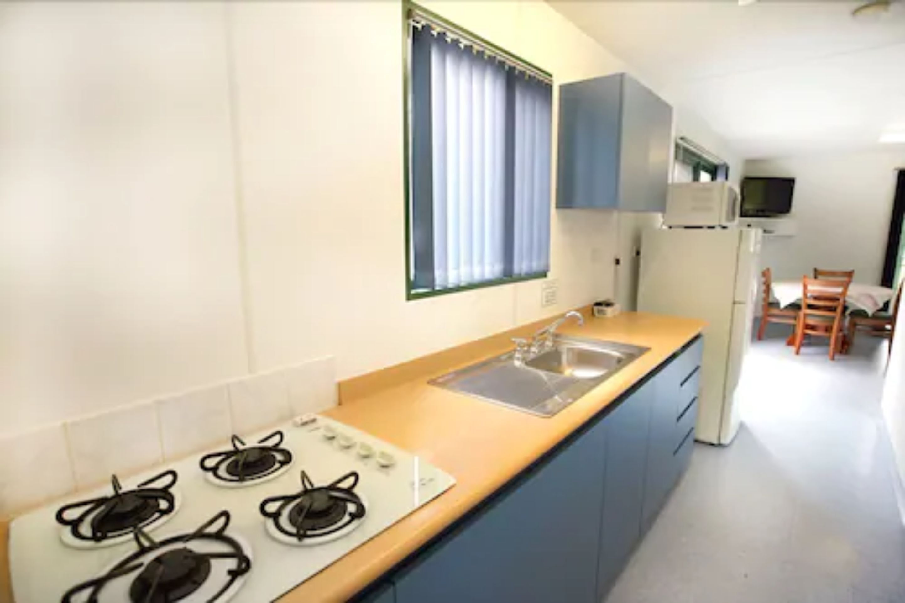 Una cocina compacta con estufa a gas, un fregadero y microondas. Hay una ventana con cortinas, un televisor montado en la pared y una zona de comedor con sillas.