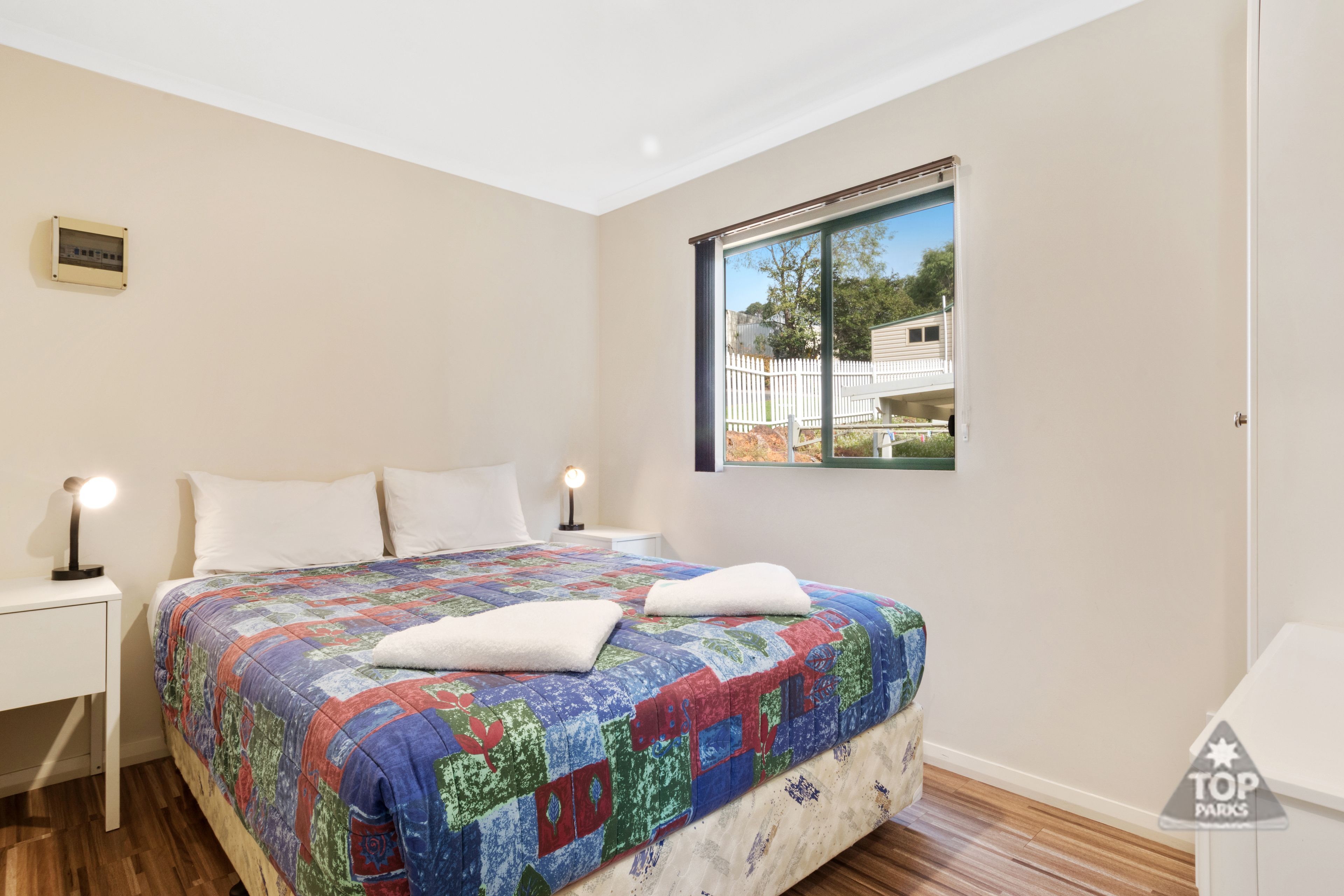 Un dormitorio con una cama grande, una mesita de noche, una lámpara de noche y una ventana con vista a una casa y una cerca.