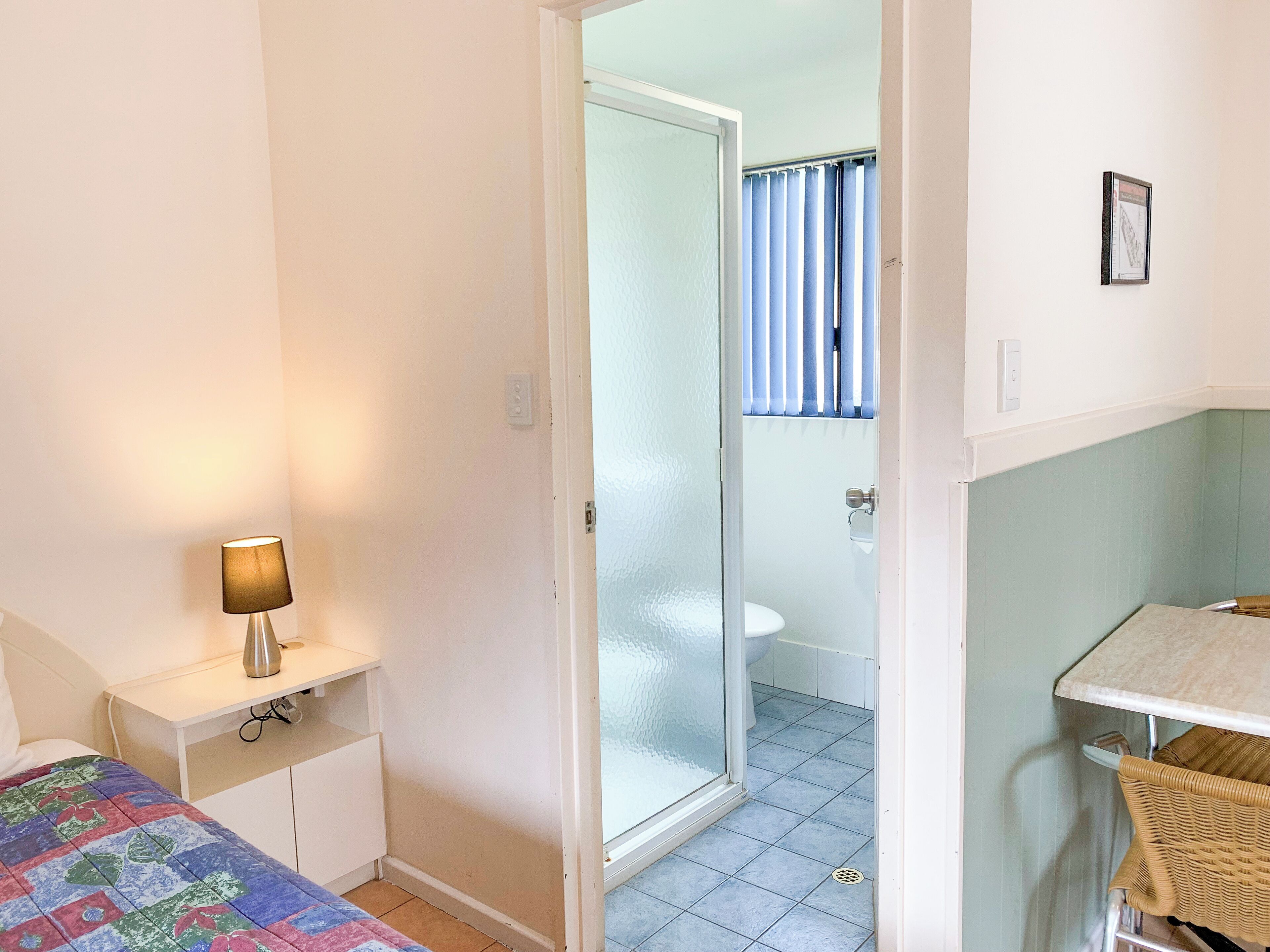 A small, well-lit room with a bed, a bedside table with a lamp, a wicker chair, and a bathroom visible through a glass door.